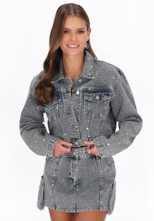 Tânără cu părul lung zâmbește, purtând o jachetă din denim gri și o fustă asortată cu buline albe, stând în fața unui fundal alb.