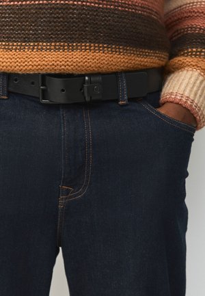 Primo piano di una persona che indossa jeans blu scuro, cintura in pelle nera e un maglione lavorato a maglia con strisce marroni, arancioni e beige, mano in tasca.