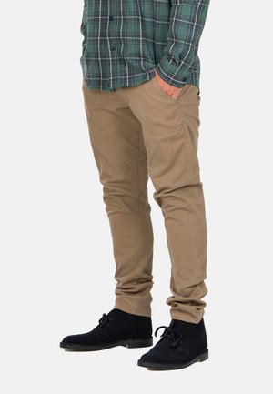 Beige Baumwollhosen mit slim fit, ausgestattet mit seitlichen Taschen, kombiniert mit schwarzen Wildleder-Ankle-Boots. Grünes Karohemd sichtbar.