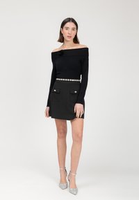 Schwarzes Off-Shoulder-Top mit Blumenakzent, kombiniert mit einem schwarzen Minirock mit zwei Fronttaschen und silbernen Knöpfen, dazu silberne Glitzer-Absätze.