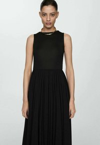 Schwarzes ärmelloses Kleid mit tailliertem Oberteil und plissiertem Rock. Glatte Textur und minimalistisches Design, ergänzt durch eine goldene Halskette.