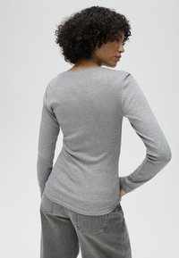 Donna con i capelli ricci che indossa una maglietta grigia aderente a maniche lunghe e jeans grigi, leggermente girata di lato con una mano sul fianco.