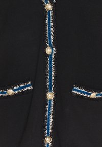 Zwart stof met gouden leeuwenkopknopen, blauwe fluwelen rand en textuur zwart-wit afwerking langs de knopenlijn en zakflappen.