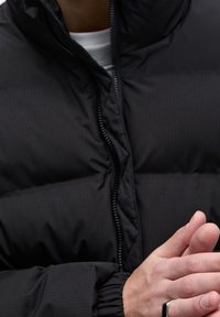 Personne portant une doudoune noire à fermeture éclair avant, joignant les mains près de la poitrine, portant une bague noire au doigt.