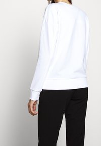 Felpa bianca in cotone con vestibilità comoda, presenta maniche lunghe e polsini a coste. Abbinata a pantaloni neri, design semplice senza motivi.