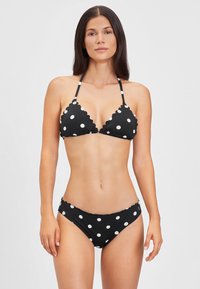 Črn bikini z belimi pikicami, ki vsebuje dizajn z valovitim robom. Zgornji del je v obliki trikotnika z tankimi naramnicami, spodnji del pa ima podoben valovit detajl.