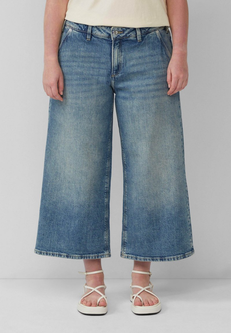 Hellblaue, weit geschnittene Jeans aus Denim mit hoher Taille, Fronttaschen und rohem Saum. Kombiniert mit weißen Riemchensandalen.