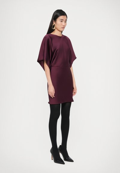 Rochie din satin de culoare marsala, cu design de mâneci largi tip capă, asortată cu colanți negri și pantofi stiletto cu vârf ascuțit. Modelul stă pe un fundal neutru.