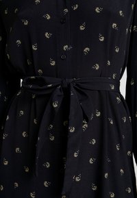 Svart klänning med knäppning fram, prydd med små gula blommönster. Inkluderar ett matchande tygbälte knutet i midjan. Mjuk textur.