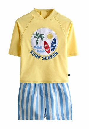 Kollane lühikeste varrukatega rash guard, millel on „Surf Seeker“ tempel, kus on palmipuu, päike, lainelauad ja sinised lained, kombineeritud siniste ja valgete triibuliste lühikeste pükstega.