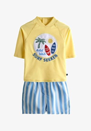 Rash guard jaune à manches courtes avec patch « Surf Seeker » représentant un palmier, le soleil, des planches de surf et des vagues bleues, associé à un short rayé bleu et blanc.