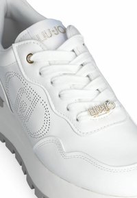 Zapatilla blanca hecha de cuero liso con detalles perforados, cordones planos blancos, acentos dorados y una suela de goma de color gris claro.