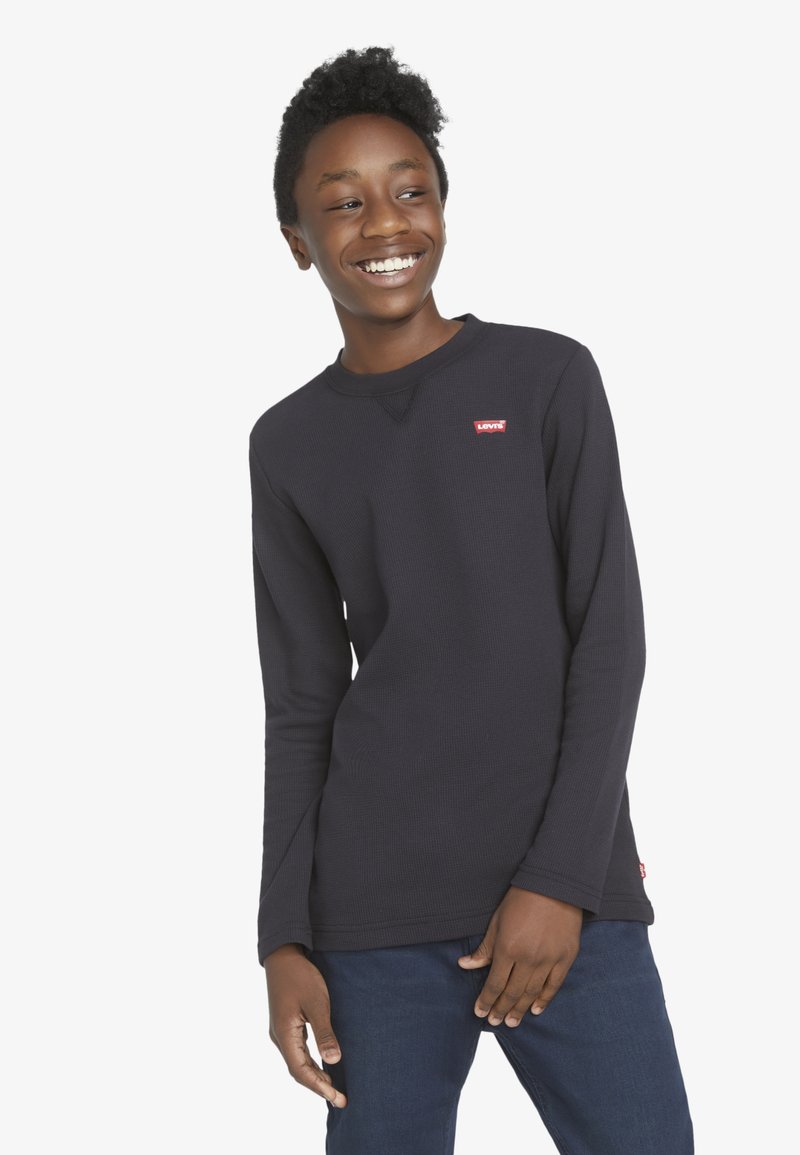 Ragazzo adolescente sorridente che guarda di lato, indossa una maglietta nera a maniche lunghe Levi's e jeans blu scuro su uno sfondo bianco.