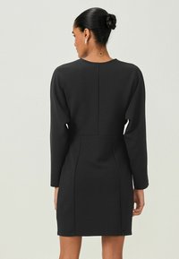 Abito nero con maniche lunghe e vita aderente, caratterizzato da una texture liscia, un design strutturato e un dettaglio della cucitura posteriore.