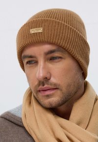 Brown ribbed knit beanie with a leather logo patch, paired with a beige scarf. The fabric has a soft, textured appearance.