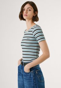 Young woman with short brown hair wearing a light blue, black, and white striped fitted shirt and blue jeans, hands in pockets, neutral background.