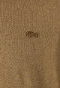 Rudas megztinis su tekstūruota paviršiumi ir siuvinėtu krokodilo logotipu kairėje krūtinės pusėje. Minkšta, lengva medžiaga.