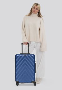 Donna con un maglione beige a collo alto e pantaloni bianchi che tiene il manico di una valigia rigida blu con ruote, su uno sfondo neutro.