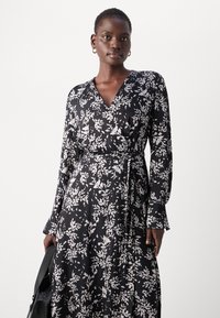 Crna haljina na preklop s bijelim cvjetnim uzorkom, dugim rukavima i remenom za vezanje. Izrađena od mekanog materijala, s dubokim V-izrezom i lepršavim siluetom.