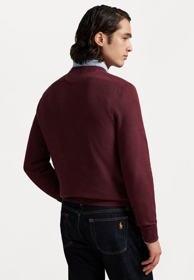 Suéter burdeos con un tejido texturizado, corte moderno, puños acanalados, llevado sobre una camisa de cuello azul claro, combinado con jeans de mezclilla oscura.