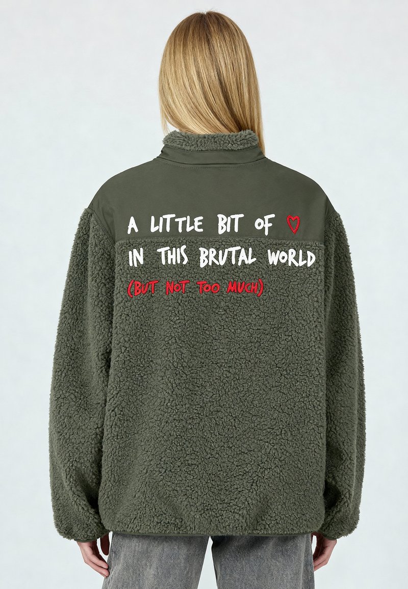 Veste en polaire vert olive avec une finition texturée, comportant un texte blanc et rouge au dos incluant un graphique de cœur.