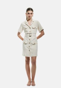 Crème korte mouwen jurk met riem, knoopsluiting, twee voorzakken en goudkleurige knopen. Glad materiaal met een getailleerde pasvorm.