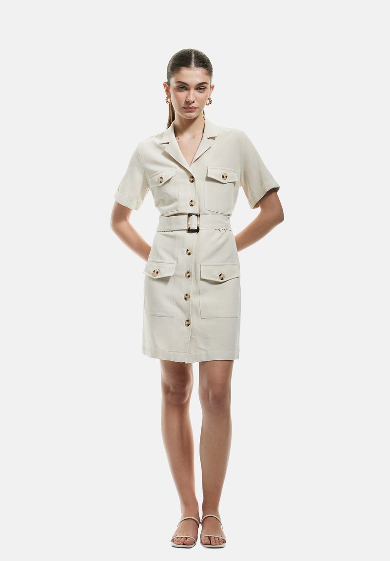 Crème korte mouwen jurk met riem, knoopsluiting, twee voorzakken en goudkleurige knopen. Glad materiaal met een getailleerde pasvorm.
