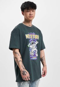 Camiseta de algodón verde oscuro con un gráfico colorido de un astronauta y el texto "SIN RETORNO". Mangas cortas y corte holgado con tatuajes visibles.