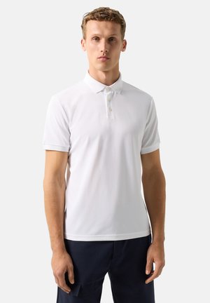 Młody mężczyzna stoi nieruchomo, ubrany w gładką białą koszulkę polo z krótkim rękawem i ciemne spodnie na neutralnym tle.