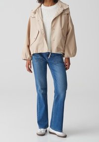 Giacca beige corta con bottoni a pressione e orlo con cordino, abbinata a jeans a zampa d'elefante blu e sneakers bianche. Sotto, un maglione crema dalla trama. 