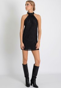 Robe mini noire à col halter avec un tissu texturé et brillant. Le modèle porte des bottes en cuir noires hautes. La robe a une coupe ajustée et un dos ouvert.