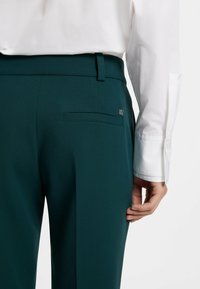 Dunkelgrüne maßgeschneiderte Hosen mit glatter Textur, featuring eine Gesäßtasche und einem kleinen metallischen Logo-Akzent am Bund.