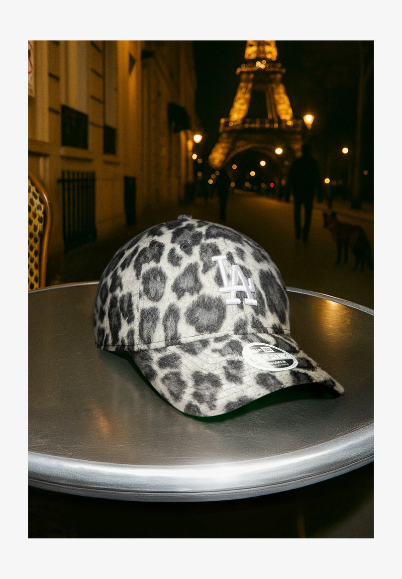 Gorra de estampado de leopardo con patrón gris y negro; cuenta con un logo blanco de LA y un visera plana. Posicionada sobre una mesa metálica.