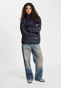 Zwarte donsjack met een hoge kraag, voorzien van een ritssluiting aan de voorkant en een doorgestikt ontwerp. Gecombineerd met lichtblauwe, loszittende jeans en grijze sneakers.