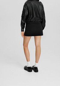 Černá bomber bunda z umělé kůže s žebrovaným lemem, v kombinaci s černou mini sukní. Černé boty a bílé ponožky dotvářejí celkový outfit.