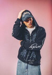 Junger Mann trägt einen schwarzen Kapuzenpullover mit Grafikdruck und eine passende Kappe mit dekorativem Text und steht vor einem pinken Hintergrund mit einer Hand am Kopf.