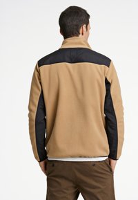 Chaqueta de forro polar en beige con detalles de nailon negro en los hombros y las mangas. Cuenta con un cuello alto y un corte relajado.