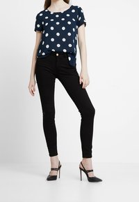 Mörkblå topp med prickar och uppvikt ärm, kombinerad med svarta slim fit-jeans och svarta remsandaler med hög klack. Slätt tyg och figurnära siluett.