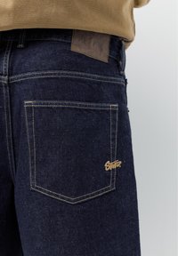 Zbliżenie na tylną kieszeń ciemnoniebieskich dżinsów z żółtym przeszyciem i małą jasnobrązową naszywką z logo przy rogu kieszeni.