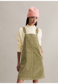 Zielona sukienka ogrodniczka z sztruksu z szerokimi ramiączkami i przednią kieszenią, zestawiona z kremowym swetrem i różową czapką beanie. Gładka tekstura, casualowy design.