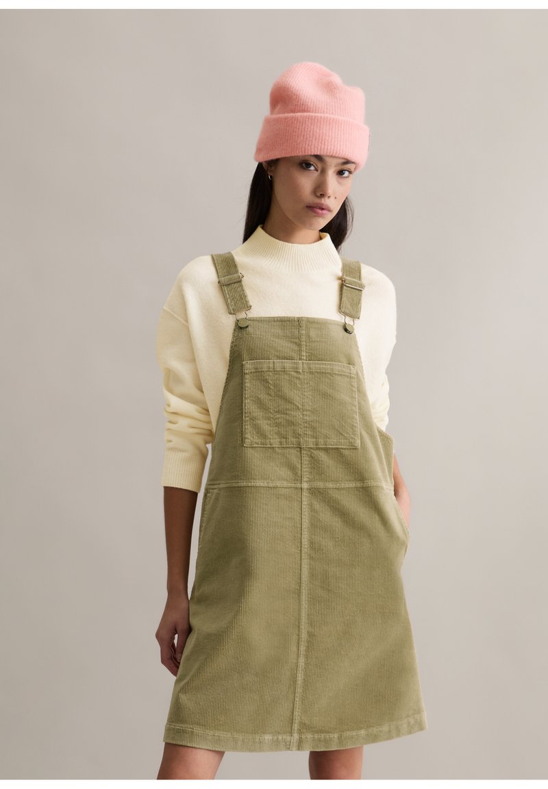 Zielona sukienka ogrodniczka z sztruksu z szerokimi ramiączkami i przednią kieszenią, zestawiona z kremowym swetrem i różową czapką beanie. Gładka tekstura, casualowy design.