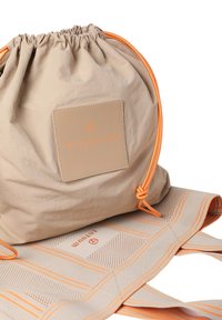 Beige trekkertje met oranje accenten en een rechthoekige, reliëf logo patch. Een gestructureerde, gemusterde mat ligt eronder.