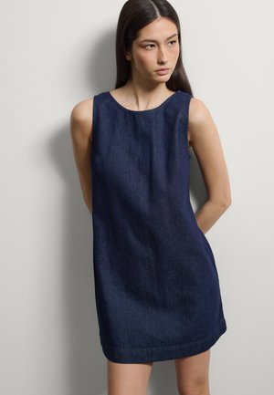 Joven mujer con cabello largo y oscuro, vestida con un vestido vaquero sin mangas de color azul oscuro, de pie contra una pared blanca lisa, mirando hacia un lado.