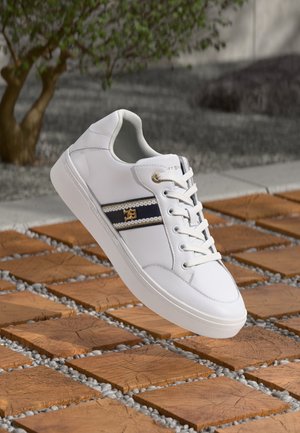 Hvid sneaker med sorte og gyldne stribedetaljer, hvide snørebånd, hvilende på træningskvadrater med grusmellemrum, sløret træ og baggrund.