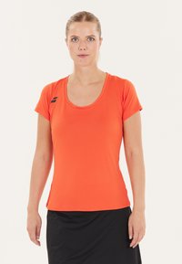 Orange kortærmet atletisk skjorte med rund hals og sort logo detalje på skulderen. Parret med en sort nederdel. Glat tekstur.