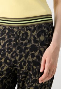 Luipaardprint broeken hebben een bruin en groen kleurenschema met een zwarte tailleband, verfraaid met twee dunne groene strepen. Soepele stofstructuur.