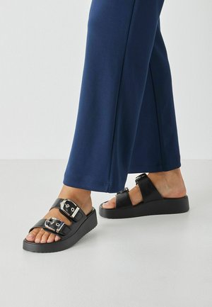 Sandales à plateforme noires avec deux larges brides, boucles métalliques et semelle en caoutchouc texturée, associées à un pantalon large bleu marine.