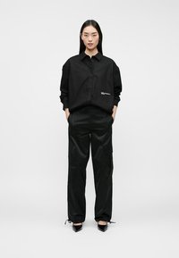 Mujer de pie, vistiendo una camisa de botones negra de gran tamaño, pantalones cargo negros y tacones de punta negros, con las manos en los bolsillos, fondo neutro.