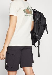 T-shirt beige con una grafica di un furgone e montagne sul retro, abbinata a pantaloni cargo neri e uno zaino nero appoggiato su una spalla.