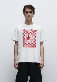 Bílá bavlněná tričko s červeným potiskem a kostkovaným okrajem, textem "Pizza Planet" a kreslenými postavičkami. Volný střih, krátké rukávy.